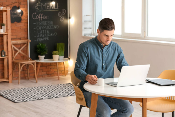 Schöner junger Mann trinkt Kaffee, während er mit Laptop im Café arbeitet - Foto, Bild