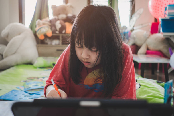 Ázsiai kis iskolás lány tanul az online leckéjét otthon mosollyal és boldog, társadalmi távolság karantén alatt, online oktatási koncepció - Fotó, kép