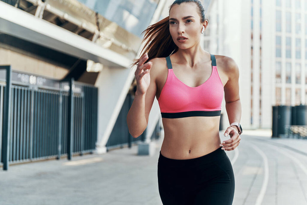Beautiful young woman in sports clothing wearing headphones while running outdoors - Foto, Bild