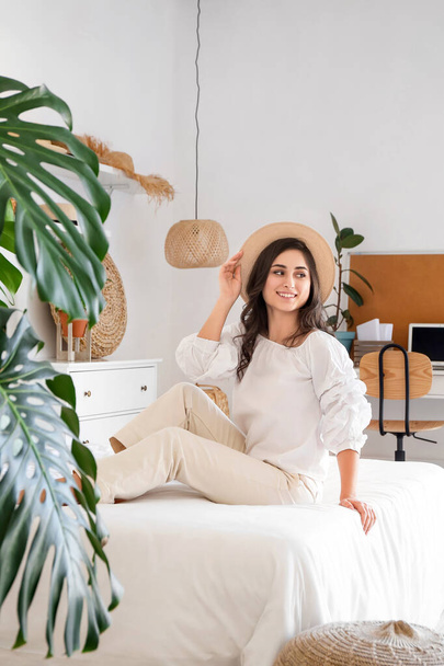 Young woman in stylish hat sitting on bed in room - Foto, Imagem