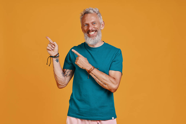 Happy senior man pointing copy space and smiling while standing against orange background - 写真・画像