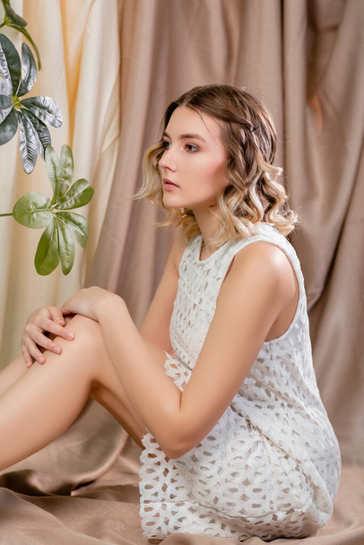 Portrait of a beautiful young woman in a white dress sitting on the floor, against the background of a light beige drapery. - Photo, image