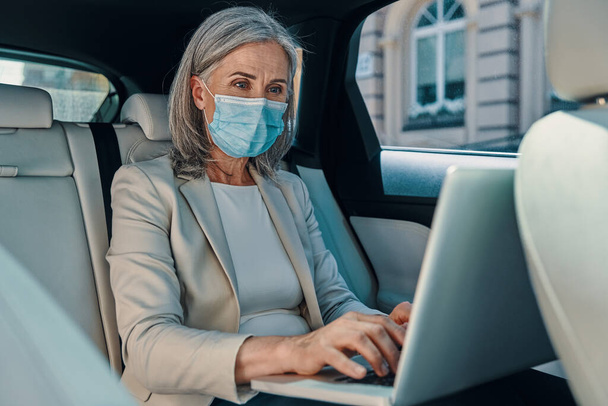 Mature beautiful woman in protective face mask using laptop while sitting on the back car seat - Zdjęcie, obraz