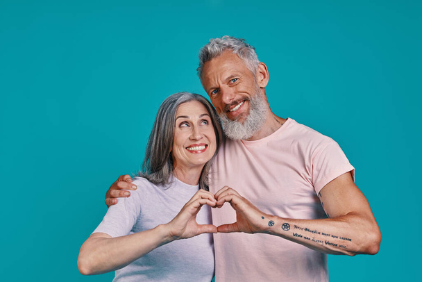 Beautiful senior couple looking at camera and showing heart with hands while standing together against blue background - Foto, Imagem