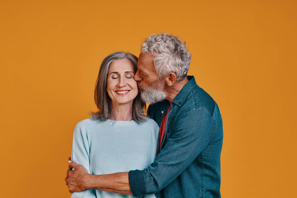 Beautiful senior couple kissing and smiling while standing together against orange background - 写真・画像