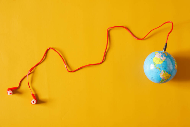 Red wired earbuds are connected to a small globe. The concept of broadcasting, webcasting, podcasting and broadcasting worldwide popular audio hits. Yellow background - Photo, image