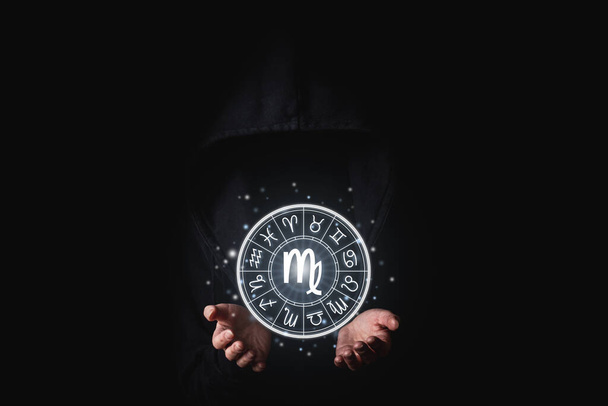 Eine Frau in schwarzem Mantel hält mit ihren Handflächen die leuchtenden astrologischen Zeichen der Tierkreiszeichen auf schwarzem Hintergrund. - Foto, Bild