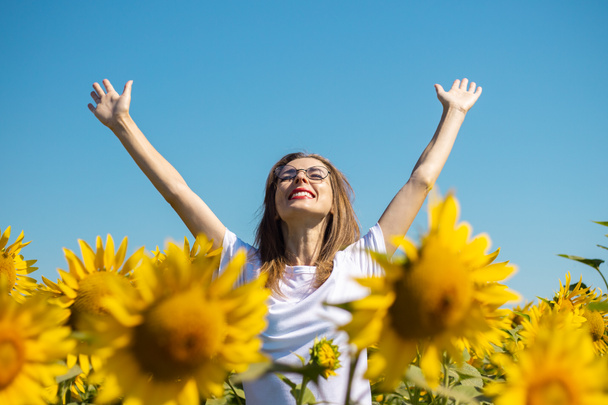 Jeune femme dans un t-shirt blanc et des lunettes avec les mains levées sur un champ de tournesol par une journée ensoleillée d'été. - Photo, image