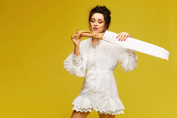 Young model woman with red manicure and bright makeup wearing white summer dress posing with crispy baguette over the yellow background - Foto, Bild
