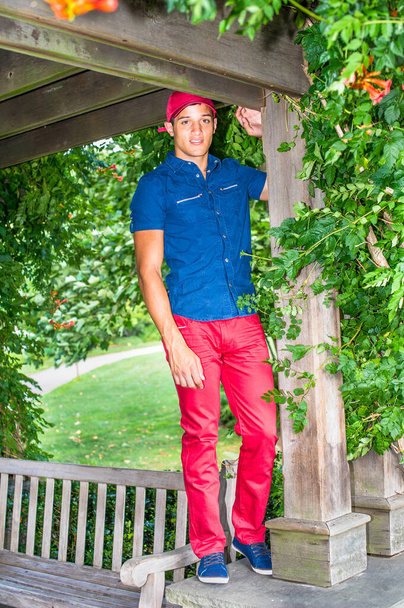 Vestirse con un sombrero de béisbol rojo, una camisa azul de manga corta, pantalones rojos y zapatos azules, un chico guapo y atractivo está jugando en el parque. - Foto, Imagen