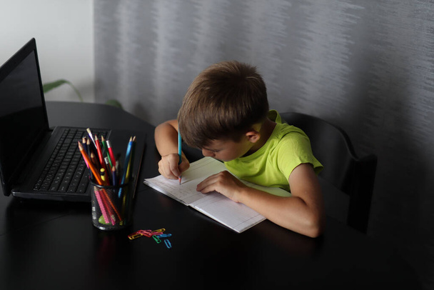 A szorgalmas fiú megtanulja a leckét, és otthon ír egy jegyzetfüzetbe. Az asztalon van egy laptop és egy kosár tollakkal. Oktatás és vissza az iskolába - Fotó, kép