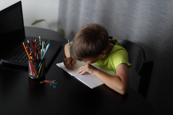 Το αγόρι γράφει εργασίες σε ένα σημειωματάριο στο σπίτι. Στο τραπέζι υπάρχει ένας φορητός υπολογιστής και ένα καλάθι με στυλό. Εκπαιδευτική ιδέα. Κλείσιμο και αντιγραφή χώρου - Φωτογραφία, εικόνα