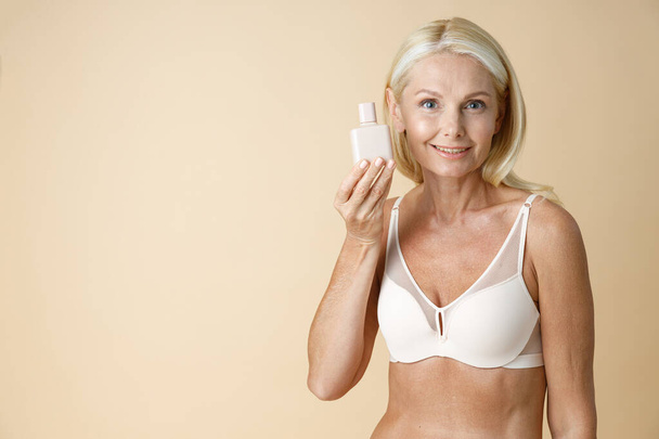 Portrait of mature blonde woman in underwear with perfect body smiling at camera, holding white bottle of cream or lotion for skin care, standing isolated over beige background - Foto, imagen
