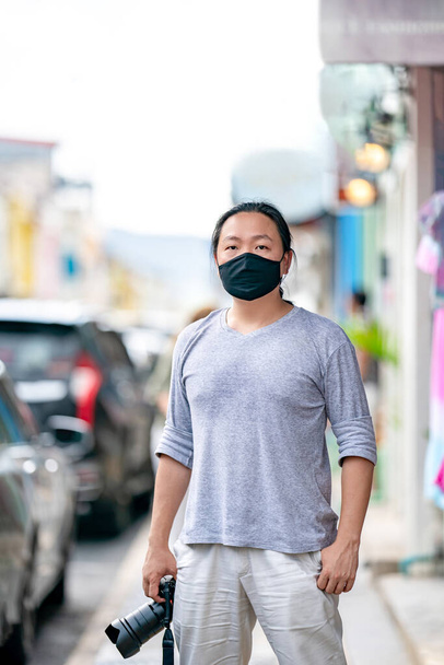 asiático profesional cámara hombre usa negro máscara y sostiene Mirrorless cámara, se encuentra en pie al lado de aparcamiento en la ciudad. - Foto, Imagen