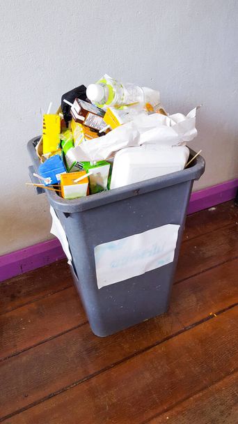 Сірий смітник на дерев'яній підлозі наповнений сміттям, яке майже виливається з смітника
  - Фото, зображення