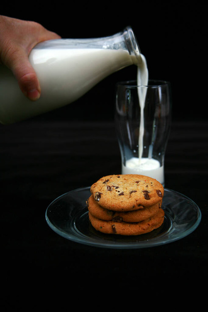 ミルクとクッキー。ミルクとチョコレートチップクッキー。1つの大きなチョコレートチップクッキーと新鮮な冷たいミルクのレトロなボトル。ミルクボトルとクッキー。昔ながらの料理やお菓子。黒ベルベットに隔離された牛乳とクッキーのガラス.  - 写真・画像