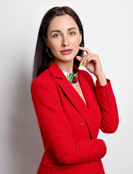 Portrait of businesswoman with permanent makeup in red jacket and modern jewelry standing sideways, holding hand at face - Fotoğraf, Görsel