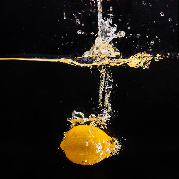 Fresh yellow lemon falling into the water with a splash of water and air bubbles, isolated on a black background. - 写真・画像