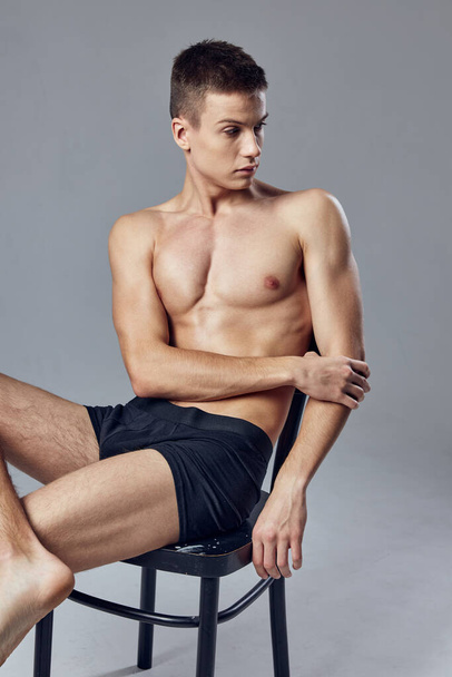 cute guy black shorts sitting on a chair look to the side studio gray background - Zdjęcie, obraz