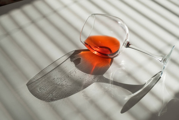 A glass of red wine lies on its side on a white table with a shade from the blinds - 写真・画像