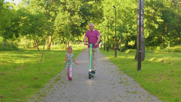 Старший современный мужчина дедушка турист верхом на электрическом скутере с внучкой девочка в парке - Фото, изображение