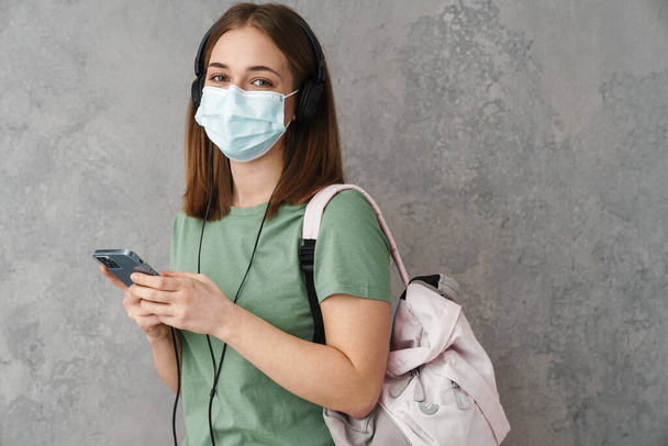 Pleased student girl in face mask using headphones and cellphone isolated over grey wall - Foto, Bild