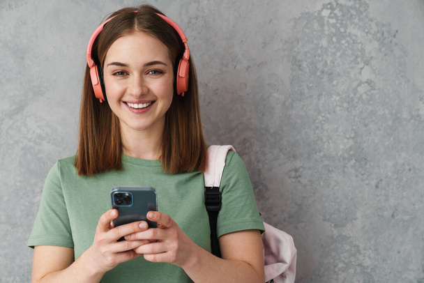 Feliz chica estudiante agradable usando auriculares y teléfono celular aislado sobre la pared gris - Foto, imagen