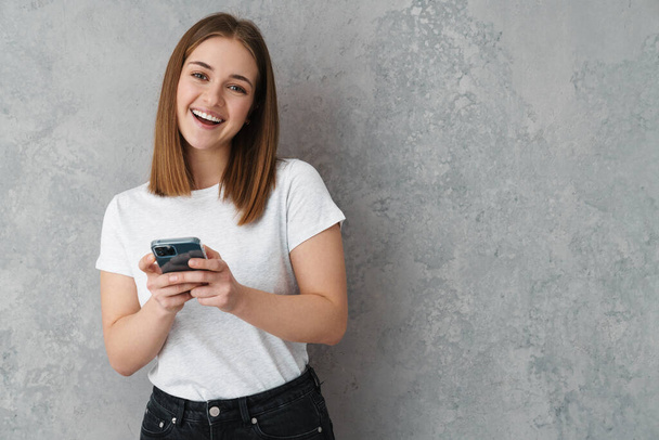 Happy nice charming girl smiling and using cellphone isolated over grey wall - Zdjęcie, obraz