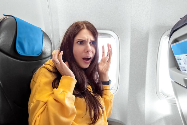 Девушка садится перед окном самолета и нервничает, боясь летать, каюту пассажирского самолета. Аэрофобия, фобия, международные рейсы, стресс, укачивание - Фото, изображение