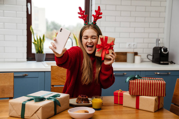 Szczęśliwa młoda dziewczyna w piżamie świętuje Boże Narodzenie przy stole w kuchni, otwiera prezenty, robi selfie - Zdjęcie, obraz
