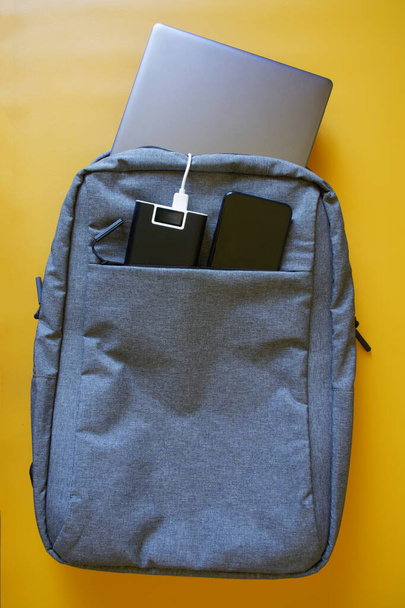 La mochila gris textil de la ciudad que contiene el portátil plateado, el smartphone y la batería exterior - el banco de fuerza sobre el fondo amarillo. Estudio, viaje y viaje con gadgets. Gradiente amarillo. Vista desde arriba - Foto, Imagen
