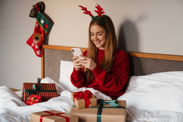 Happy young woman wearing red pajamas laying in bed with christmas presents, using mobile phone - Foto, Imagem