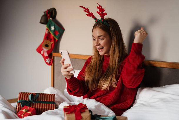 Happy young woman wearing red pajamas laying in bed with christmas presents, using mobile phone, celebrating - Foto, Imagen