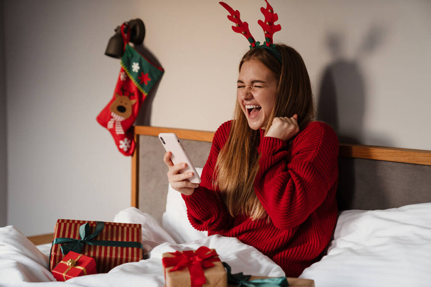 Happy young woman wearing red pajamas laying in bed with christmas presents, using mobile phone, celebrating - Photo, image