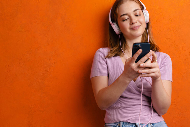 Young ginger happy woman listening music with headphones and cellphone isolated over orange wall - Foto, Imagem