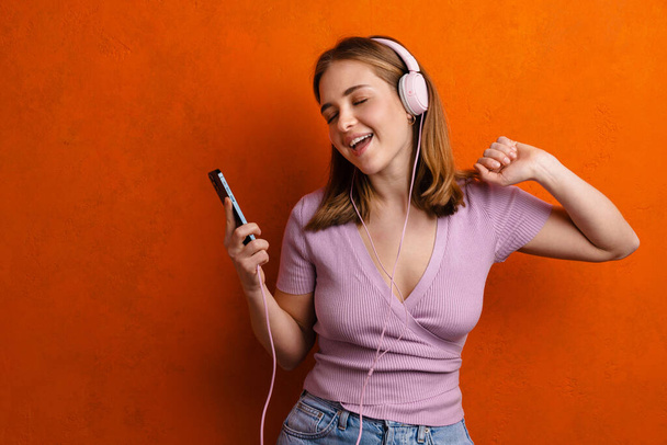 Joyful woman dancing while listening music with headphones and cellphone isolated over orange wall - Foto, Imagem
