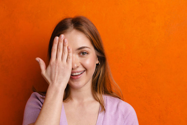 Young ginger joyful woman smiling while covering her eye isolated over orange wall - 写真・画像