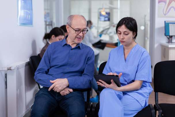 Krankenschwester macht sich Notizen am Tablet-PC und diskutiert mit Seniorin in Zahnarztpraxis - Foto, Bild