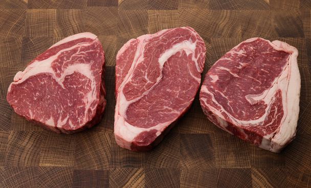 Close up three raw marbled ribeye beef steaks over end grain cutting board of wooden butcher block, high angle view, directly above - 写真・画像