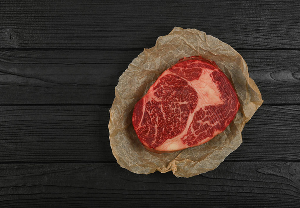 Close up one aged prime marbled raw ribeye beef steak on brown paper parchment wrapping over black wooden table background, elevated top view, directly above - Photo, image
