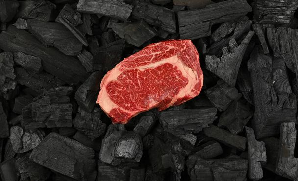 Закройте один старый мраморный сырой стейк из говядины рибай на черных кусочках древесного угля, готовый к приготовлению гриля на гриле, возвышенный вид сверху, прямо над - Фото, изображение