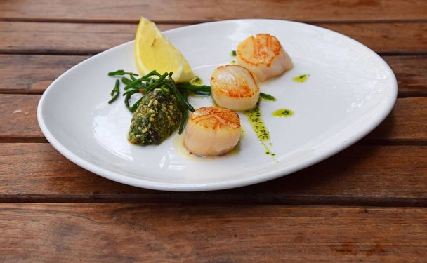 Close up portion of three grilled scallops on white plate over brown wooden table, high angle view - Photo, Image