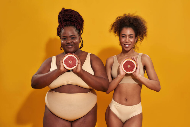 Two young african american women in underwear smiling at camera, holding halfs of juicy grapefruit in front of them, posing isolated over orange background - Photo, image