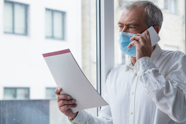 mature man with medical mask using mobile phone and flipping through documents in office, new normal concept - Фото, изображение