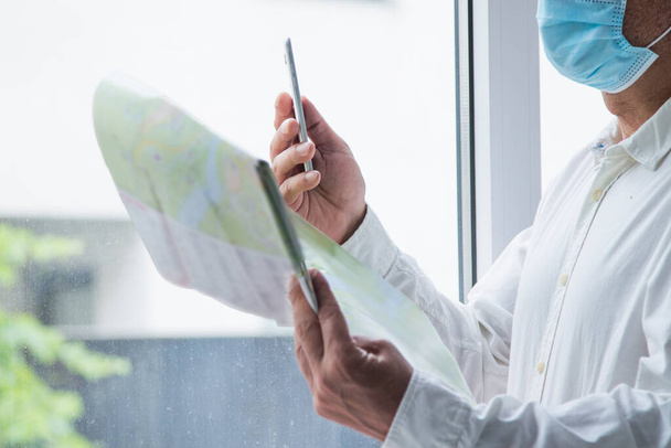 man in medical mask with a world map in his hand and in the other the mobile phone, concept of travel in the new normal - Foto, Imagen
