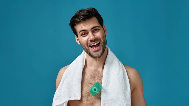 Portrait of funny brunette guy smiling at camera, holding dental floss in his mouth, posing isolated over blue background - Zdjęcie, obraz