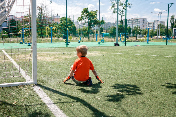 Μικρό αγόρι σε μαύρο και πορτοκαλί μορφή ποδοσφαίρου που παίζει ποδόσφαιρο σε ανοιχτό γήπεδο στην αυλή, νεαρός τερματοφύλακας - Φωτογραφία, εικόνα