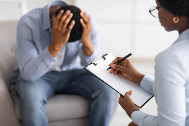 Depressieve zwarte man houdt zijn hoofd bij counselors kantoor, op zoek naar professionele hulp met psychische stoornis - Foto, afbeelding