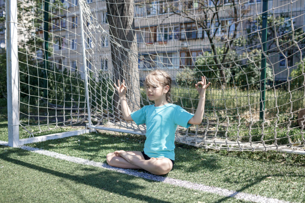 Preteen κορίτσι κάθεται στη θέση του λωτού στο τέρμα θέση στο γήπεδο, μελωδίες για να κερδίσει, προσεύχεται για την ομάδα ποδοσφαίρου - Φωτογραφία, εικόνα