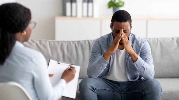 Отчаянный чёрный мужчина, страдающий неврозом или депрессией, у него сеанс в офисе психолога, панорама - Фото, изображение
